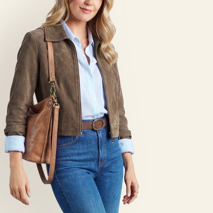 A woman modeling a brown suede jacket, blue jeans, a light blue shirt, and brown heels, carrying a brown leather shoulder bag against a solid light beige background.