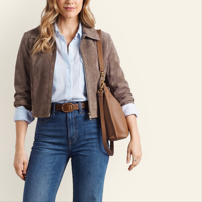 A woman wearing a light blue long-sleeve button-up shirt with chest pockets, blue jeans, a brown belt, and carrying a brown leather shoulder bag, posed against a neutral light gray background.
