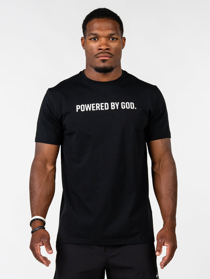 A man wearing a black short-sleeve t-shirt with the white printed text 'POWERED BY GOD.' across the chest, standing against a light gray studio background.