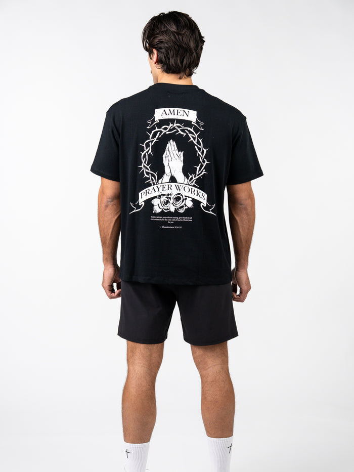 Man wearing a black Prayer Works Premium Oversized Tee featuring a white graphic of praying hands inside a crown of thorns with text banners, paired with black shorts and white socks.