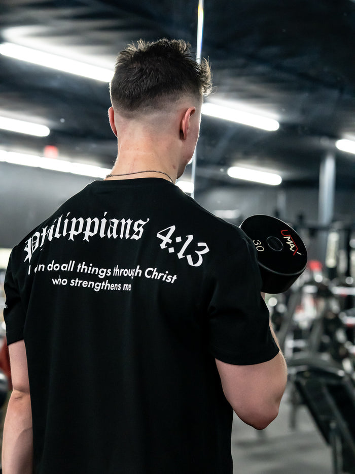 Person holding dumbbells wearing a black t-shirt with 'Philippians 4:13' text in a gym.