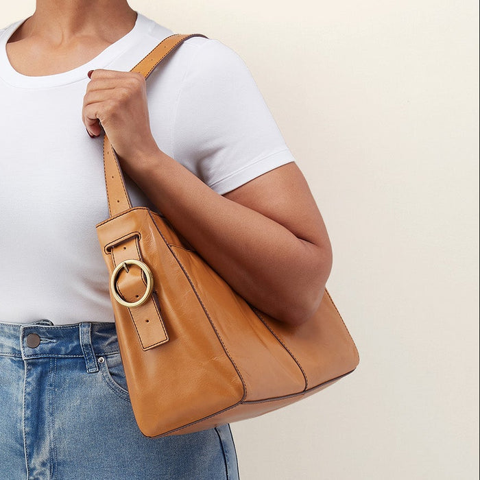 A woman wearing a white t-shirt and light blue jeans carries a tan leather shoulder bag with a large circular gold-tone ring detail on the front, set against a plain beige background.