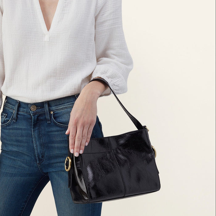 A model wearing a white long-sleeve blouse and blue jeans holds a black leather shoulder bag with gold-tone hardware against a plain light beige background.
