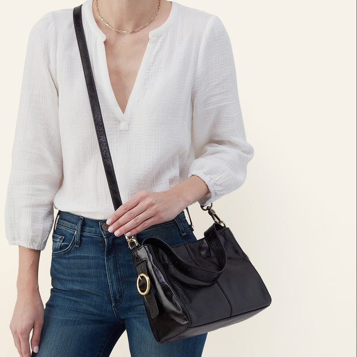 A model wearing a white blouse and blue jeans carries a black leather shoulder bag with a gold-tone circular ring detail and a black strap against a light beige background.
