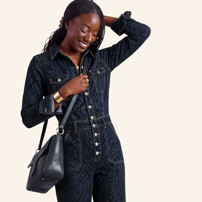 A woman modeling a dark indigo denim one-piece jumpsuit with a subtle tonal pattern, featuring gold-tone buttons down the front and side pockets. She is holding a black leather shoulder bag against a solid light beige background.