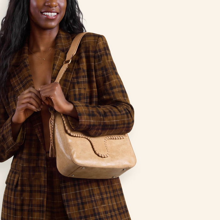 A woman modeling a brown and black plaid blazer and matching wide-leg trousers, carrying a beige shoulder bag against a solid cream background.