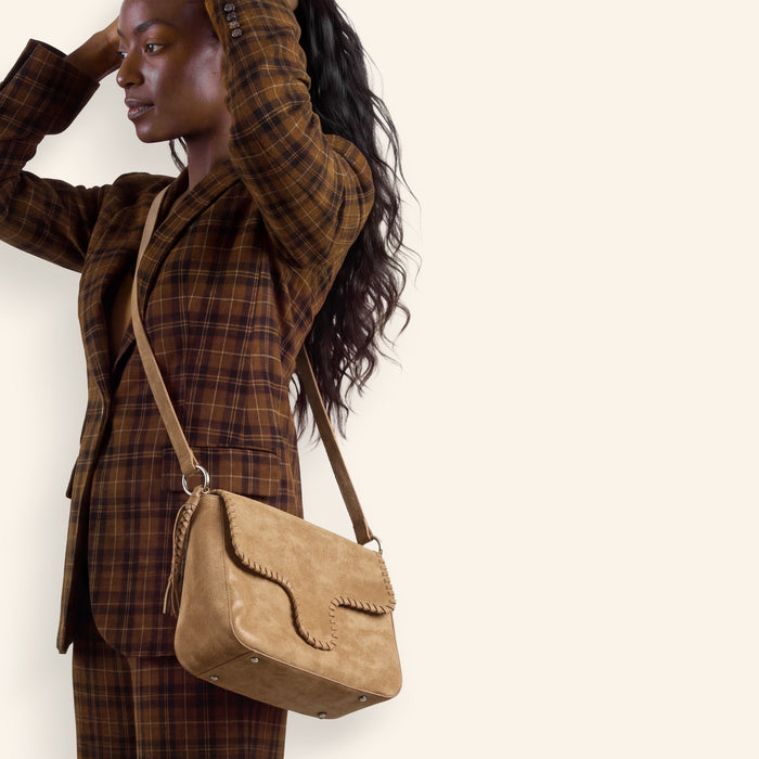 A woman modeling a brown and black plaid blazer and matching wide-leg trousers, carrying a tan suede crossbody bag, against a solid cream background.