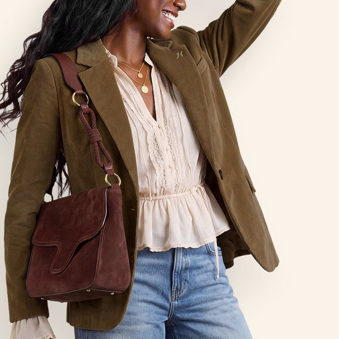 A woman posing in a studio setting wearing an olive green blazer, cream ruffled blouse, blue jeans, and carrying a brown leather shoulder bag.