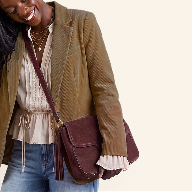A woman modeling a brown suede blazer, wide-leg blue jeans, a beige ruffled top, and a dark burgundy crossbody bag against a solid cream background.