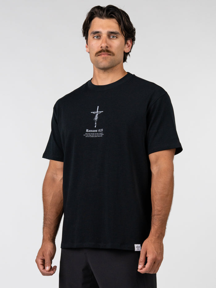 Man wearing a black Romans 6:23 “Wages of Sin” Premium Oversized Tee featuring a small cross graphic and faith-based text, styled with a relaxed oversized fit.