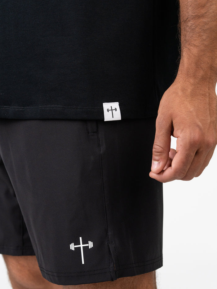 Close-up of a person wearing the Romans 6:23 “Wages of Sin” Premium Oversized Tee in black, paired with black shorts featuring a white cross and barbell logo, highlighting Christian fitness apparel.