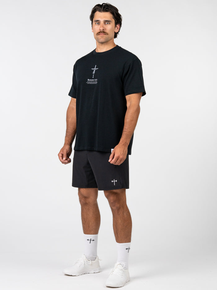 Man wearing Romans 6:23 “Wages of Sin” Premium Oversized Tee in black with a cross graphic, paired with black shorts, white socks, and white athletic shoes, showcasing Christian fitness apparel.