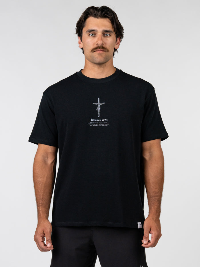Man wearing Romans 6:23 “Wages of Sin” Premium Oversized Tee in black, featuring a small crucifix graphic and scripture text, styled for Christian gym clothing and faith based apparel.