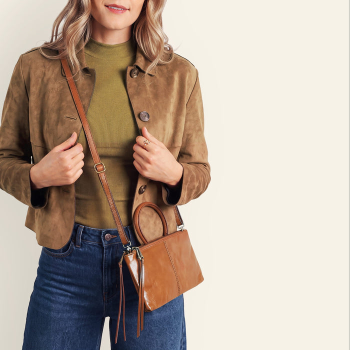 A woman modeling a brown suede jacket, olive green turtleneck, blue jeans, and a brown leather crossbody bag against a plain off-white background.