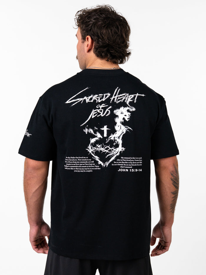 Man wearing black Sacred Heart Premium Oversized Tee with white graphic of a crowned heart, cross, barbell design, and biblical text on the back, styled as Christian fitness apparel.