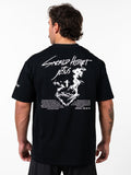 Man wearing black Sacred Heart Premium Oversized Tee with white graphic of a crowned heart, cross, barbell design, and biblical text on the back, styled as Christian fitness apparel.