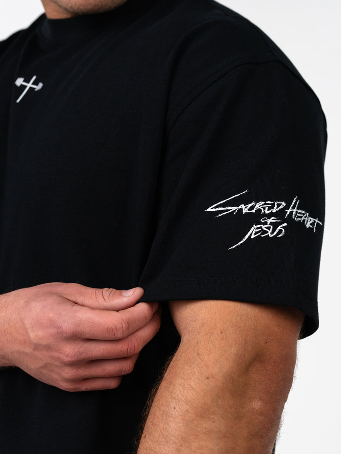 Close-up of a person wearing the Sacred Heart Premium Oversized Tee in black, featuring a small white barbell cross logo on the chest and "Sacred Heart of Jesus" text embroidered on the sleeve.