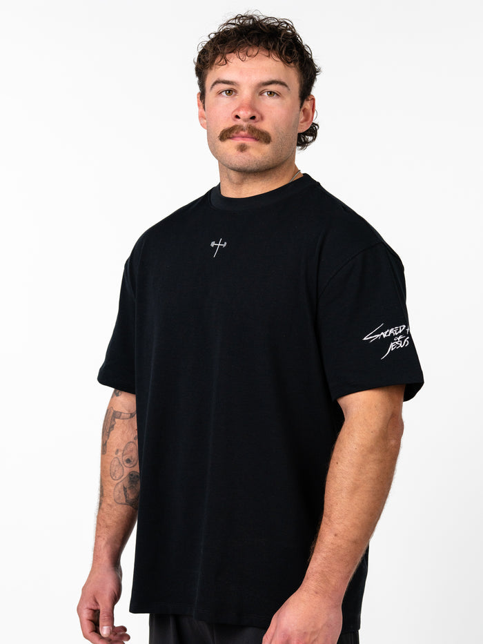 Man wearing a black Sacred Heart Premium Oversized Tee with a small cross and barbell design on the chest and "Sacred Jesus" text on the sleeve, showcasing faith based fitness apparel.