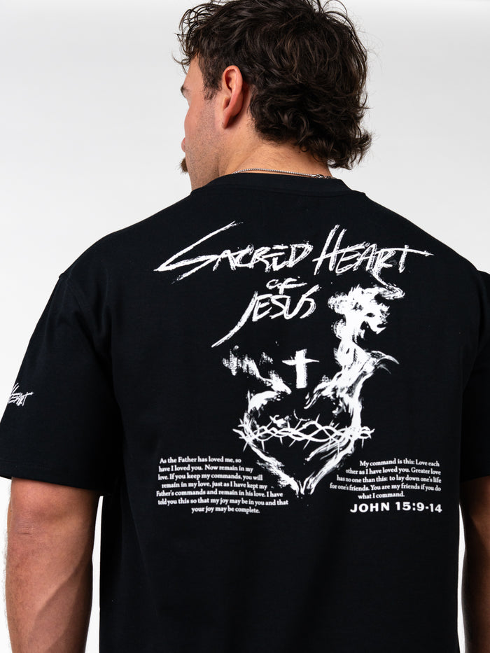 Man wearing a black Sacred Heart Premium Oversized Tee with white graphic design on the back featuring a crowned heart, cross, crown of thorns, and scripture text from John 15:9-14 in a bold, artistic style.