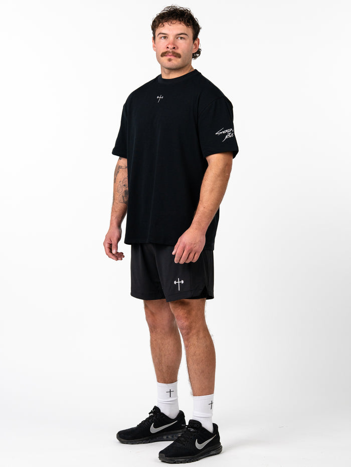 Man wearing a black Sacred Heart Premium Oversized Tee with a small cross design on the front, paired with black athletic shorts and white socks featuring a cross, styled as Christian fitness apparel.