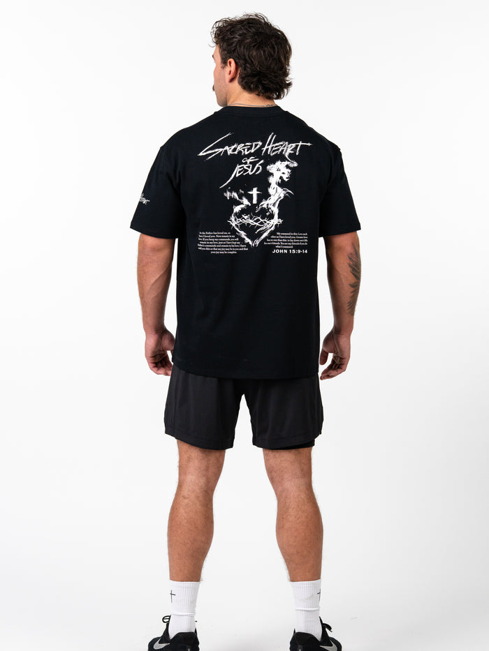 Man wearing a black Sacred Heart Premium Oversized Tee featuring white graphic text and religious imagery on the back, paired with black shorts and athletic shoes.