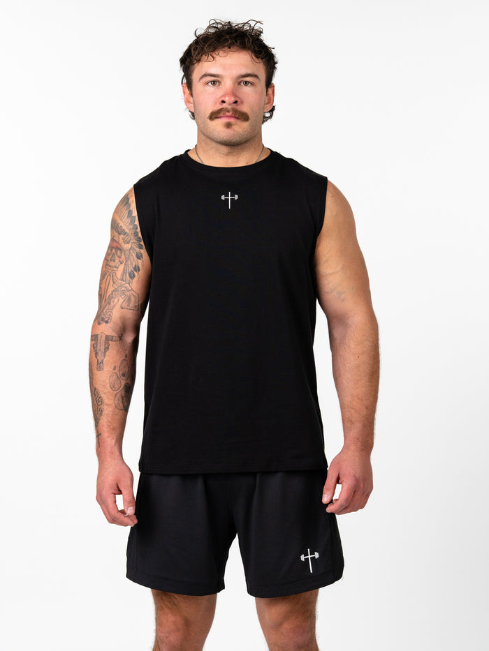 Male model wearing black Sacred Heart Premium Sleeveless Tank with a small cross and barbell design on the chest, paired with black athletic shorts featuring the same emblem.