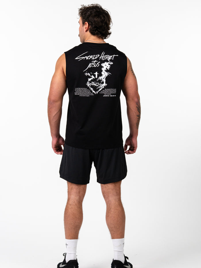 Man wearing black Sacred Heart Premium Sleeveless Tank with white graphic design featuring a sacred heart, cross, and scripture text on the back, paired with black shorts and athletic shoes.