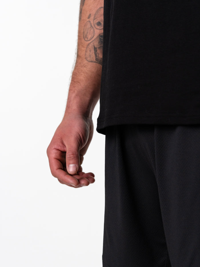 Close-up of a person's arm and hand wearing a black Sacred Heart Premium Sleeveless Tank, showcasing casual black athletic shorts and a detailed tattoo on the forearm against a white background.
