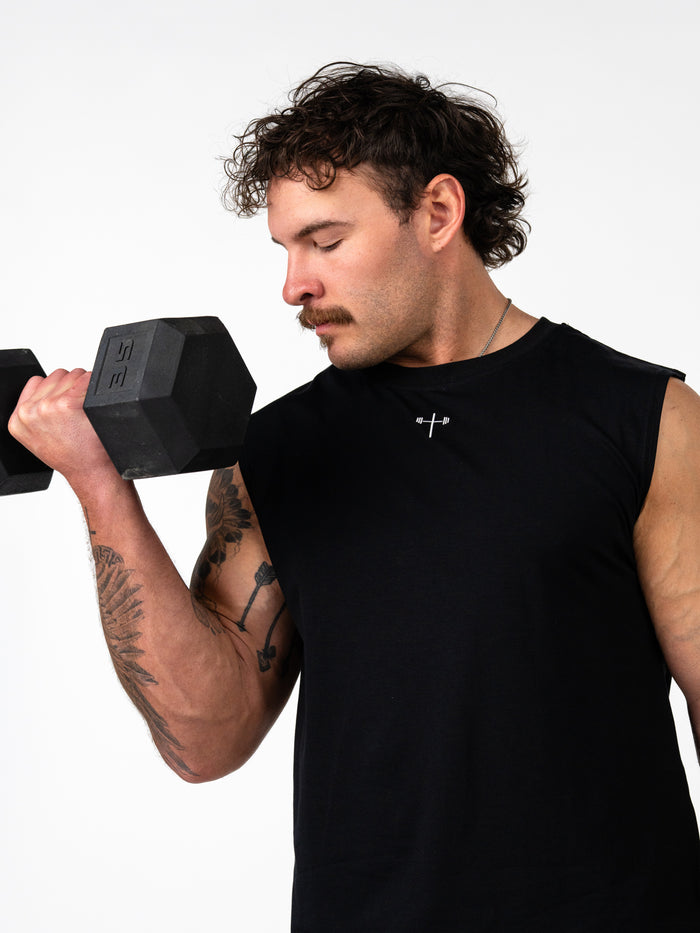 Man lifting a dumbbell while wearing a black Scripture Premium Sleeveless Tank featuring a small cross with barbell design, representing Christian fitness apparel.