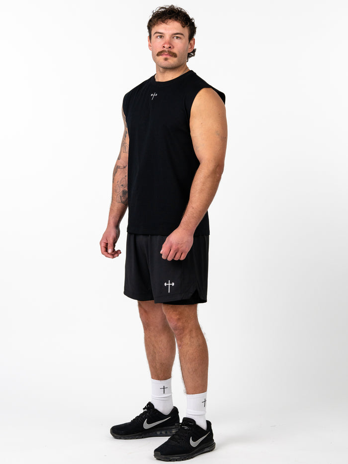 A muscular man wearing the Scripture Premium Sleeveless Tank in black, paired with matching black shorts, white socks with cross symbols, and black athletic shoes, standing against a white background.