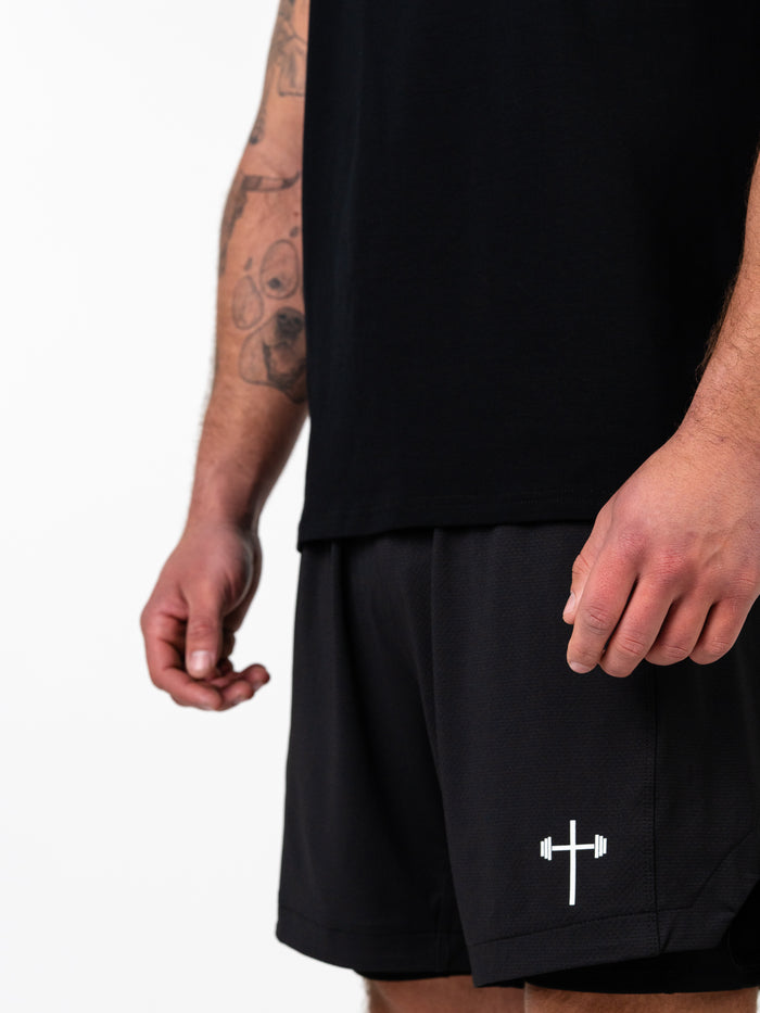 Close-up of a man wearing black shorts with a white barbell cross logo, paired with a black sleeveless top, showcasing the Scripture Premium Sleeveless Tank as part of Christian fitness apparel.