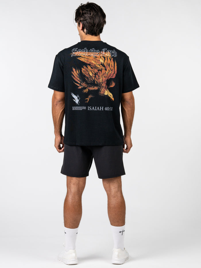 Man wearing a black Seek the Lord Premium Oversized Tee with a large graphic of a soaring eagle and script text on the back, paired with black shorts and white socks and sneakers.