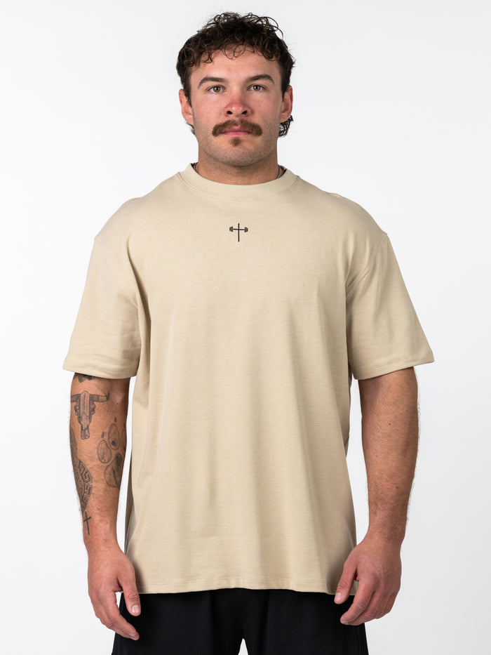 Man wearing a beige Servant of God Premium Oversized Tee featuring a small cross with a barbell design on the chest, paired with black shorts, standing against a white background.