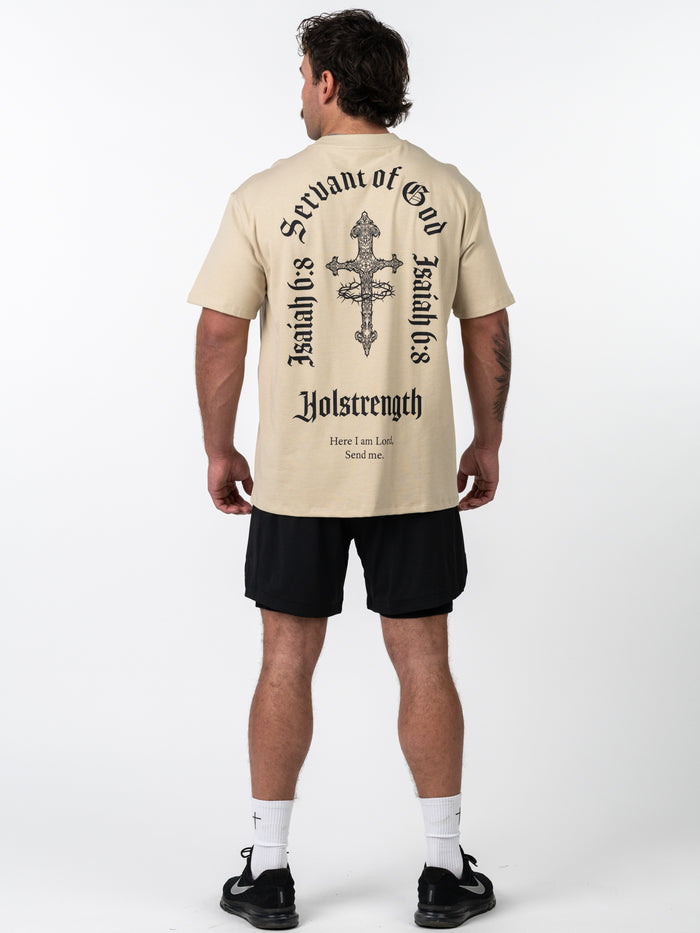 Man wearing a beige Servant of God Premium Oversized Tee with a large black cross and Isaiah 6:8 scripture on the back, paired with black shorts and athletic shoes.