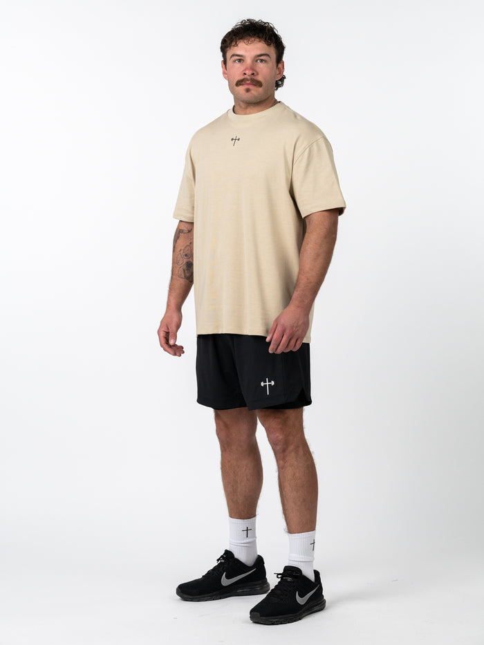 Male model wearing a beige Servant of God Premium Oversized Tee paired with black athletic shorts featuring a subtle Christian barbell cross design, styled with white socks and black Nike sneakers.