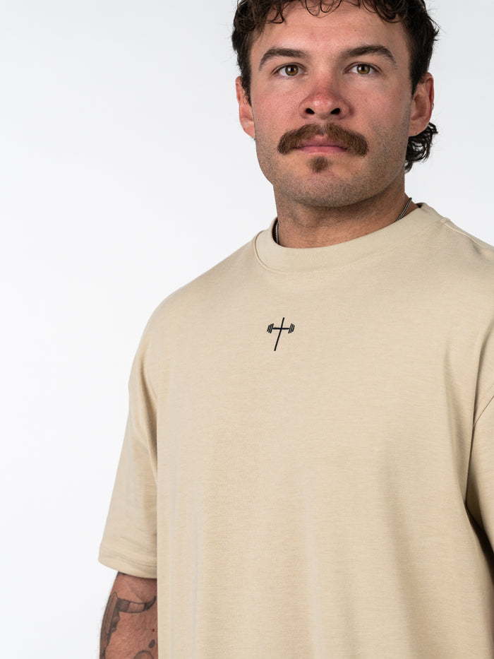 Close-up of a man wearing a beige Servant of God Premium Oversized Tee featuring a small black barbell cross emblem on the chest, blending Christian fitness apparel with casual style.