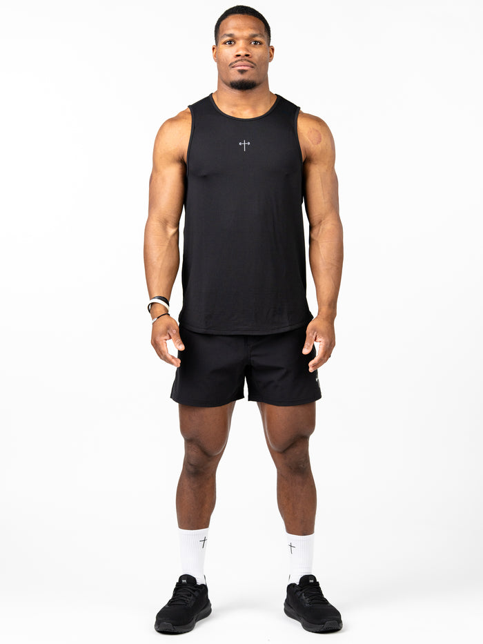 Man wearing a black Signature Tank with a small cross design on the chest, paired with black shorts, white socks featuring a cross, and black athletic shoes, standing against a white background.