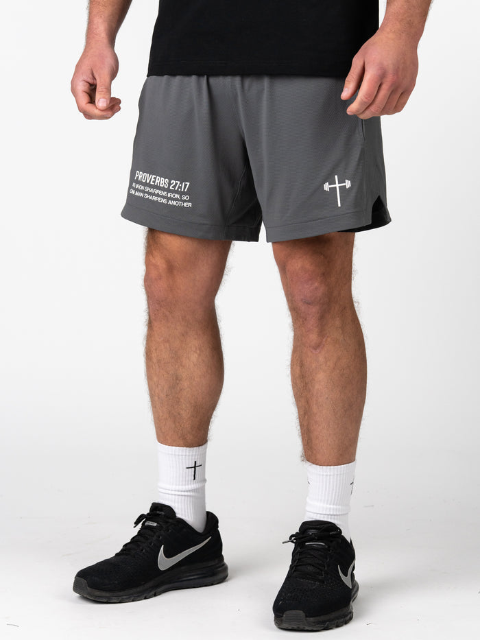 Man wearing gray Signature-Tech Liner Shorts featuring a Christian barbell cross logo and Proverbs 27:17 scripture on the left leg, paired with white socks and black Nike sneakers for training.