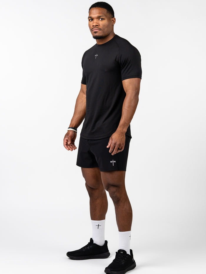 Athletic man wearing a black Signature Tee made from breathable, sweat-wicking fabric with a subtle Christian cross and barbell design, paired with matching shorts and white socks.