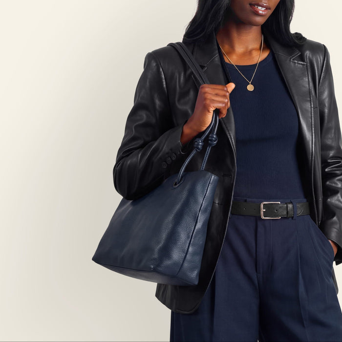 A model wearing a black leather blazer, navy blue top and wide-leg trousers, a navy cap, and holding a navy leather tote bag against a light beige background.