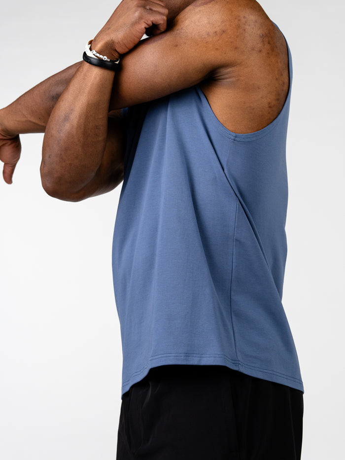 Man stretching arm wearing a blue Vital Cross Sleeveless Tank designed for high-output training and unrestricted movement, paired with black shorts against a white background.