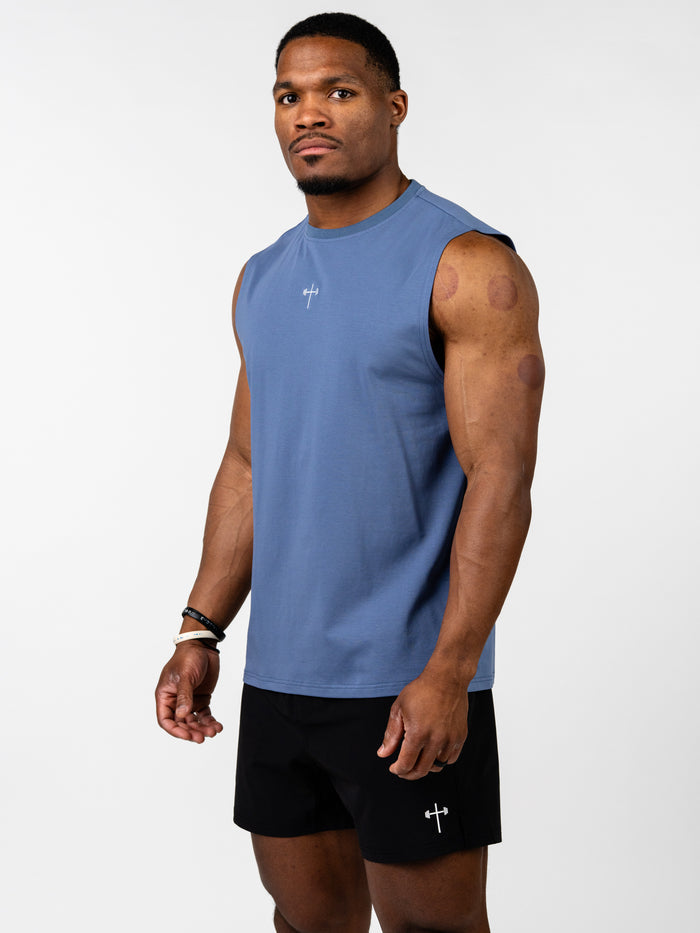 Athletic man wearing a blue Vital Cross Sleeveless Tank designed for high-output training, paired with black shorts featuring a small cross and barbell logo, standing against a plain background.