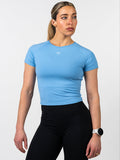 Woman wearing a light blue Women's Combat Tee with a small white cross and barbell design on the chest, paired with black workout leggings and a white fitness watch.