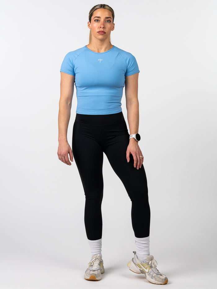 Woman wearing a fitted light blue Women's Combat Tee with a small white Christian cross and barbell graphic on the chest, paired with black leggings and athletic shoes.