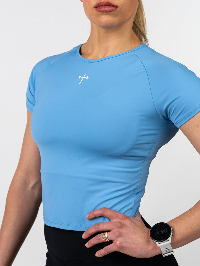 Woman wearing a blue Women's Combat Tee with a small white barbell cross logo on the chest, paired with black activewear bottoms and a smartwatch, showcasing fitted performance apparel.