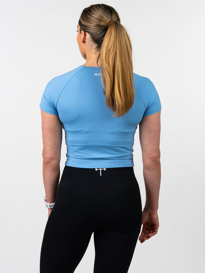 Woman wearing a fitted light blue Women's Combat Tee designed for performance, paired with black leggings, viewed from the back, standing against a plain white background.