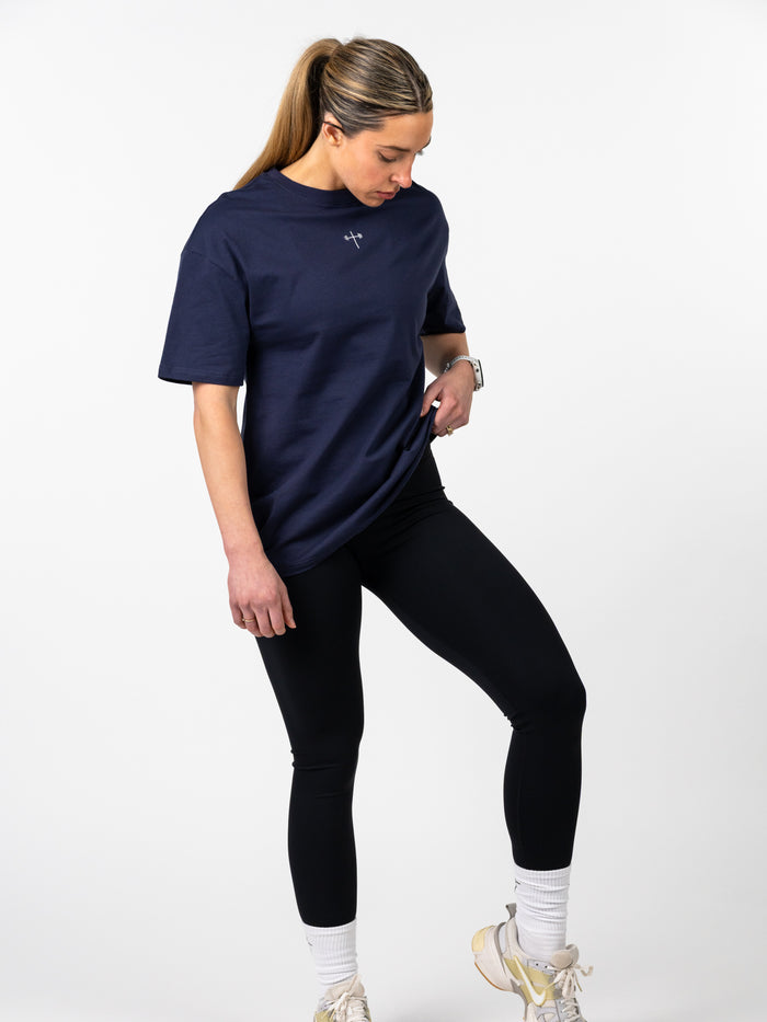 Woman wearing a navy Women's Lifting Club Oversized Tee with a small subtle Christian cross and barbell design on the chest, paired with black leggings and athletic shoes.