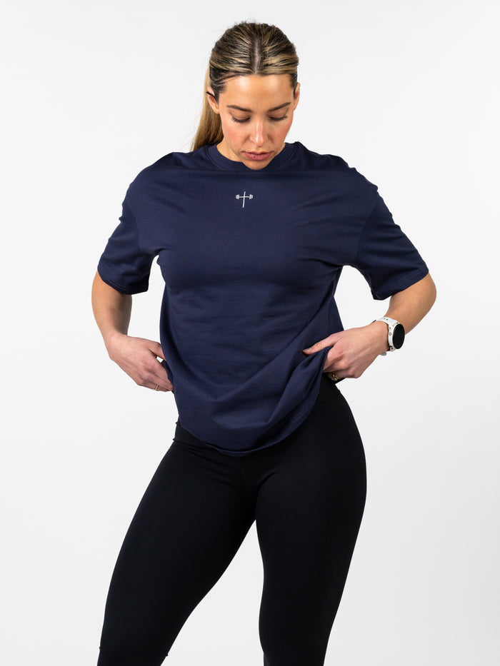 Woman wearing a navy Women's Lifting Club Oversized Tee featuring a small barbell cross design on the chest, styled with black leggings and a fitness watch.