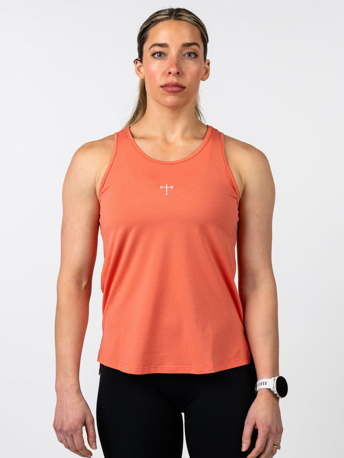 Woman wearing a coral Women's Signature Tank featuring a small white cross with a barbell design on the chest, paired with black leggings and a white fitness watch.