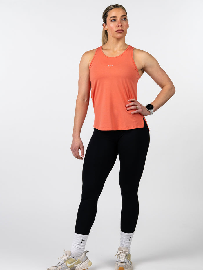 Woman wearing an orange Women's Signature Tank made from soft cotton/spandex blend, paired with black leggings and white socks with a subtle cross design, standing confidently.